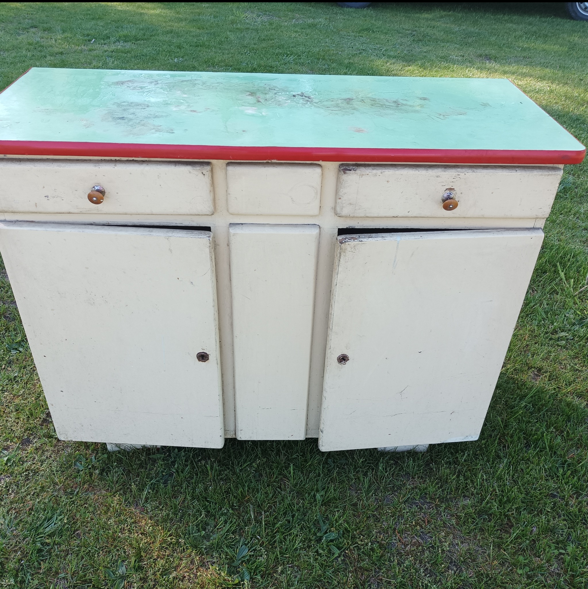 Stara, odrestaurowana szafka z zielonym blatem i czerwonym obramowaniem, stojąca na trawie, z otwartymi drzwiczkami ukazującymi wnętrze.