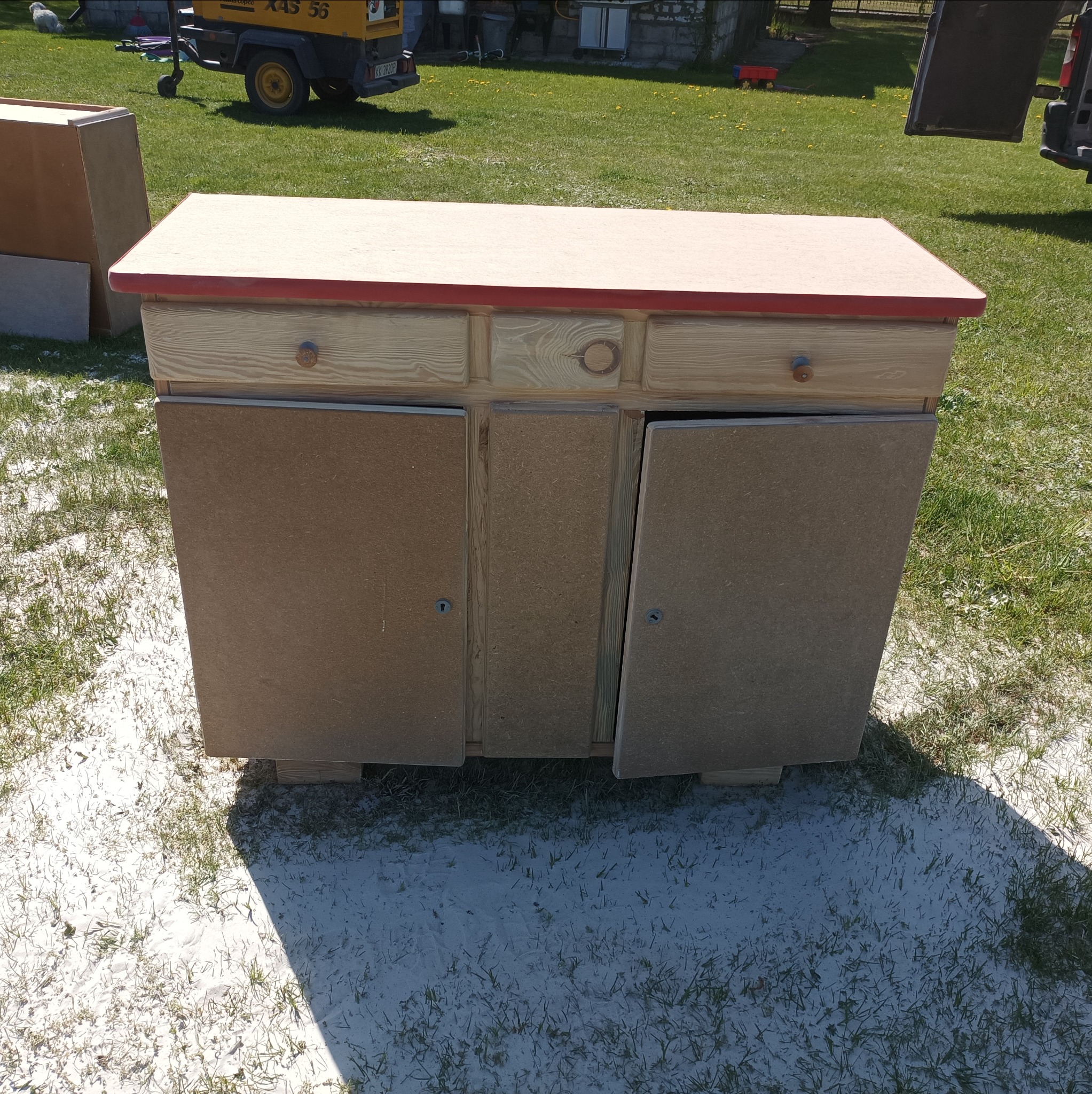 Drewniana komoda z otwartymi drzwiczkami, stojąca na trawie pokrytej białym pyłem, prawdopodobnie po piaskowaniu, z widoczną sprężarką w tle.