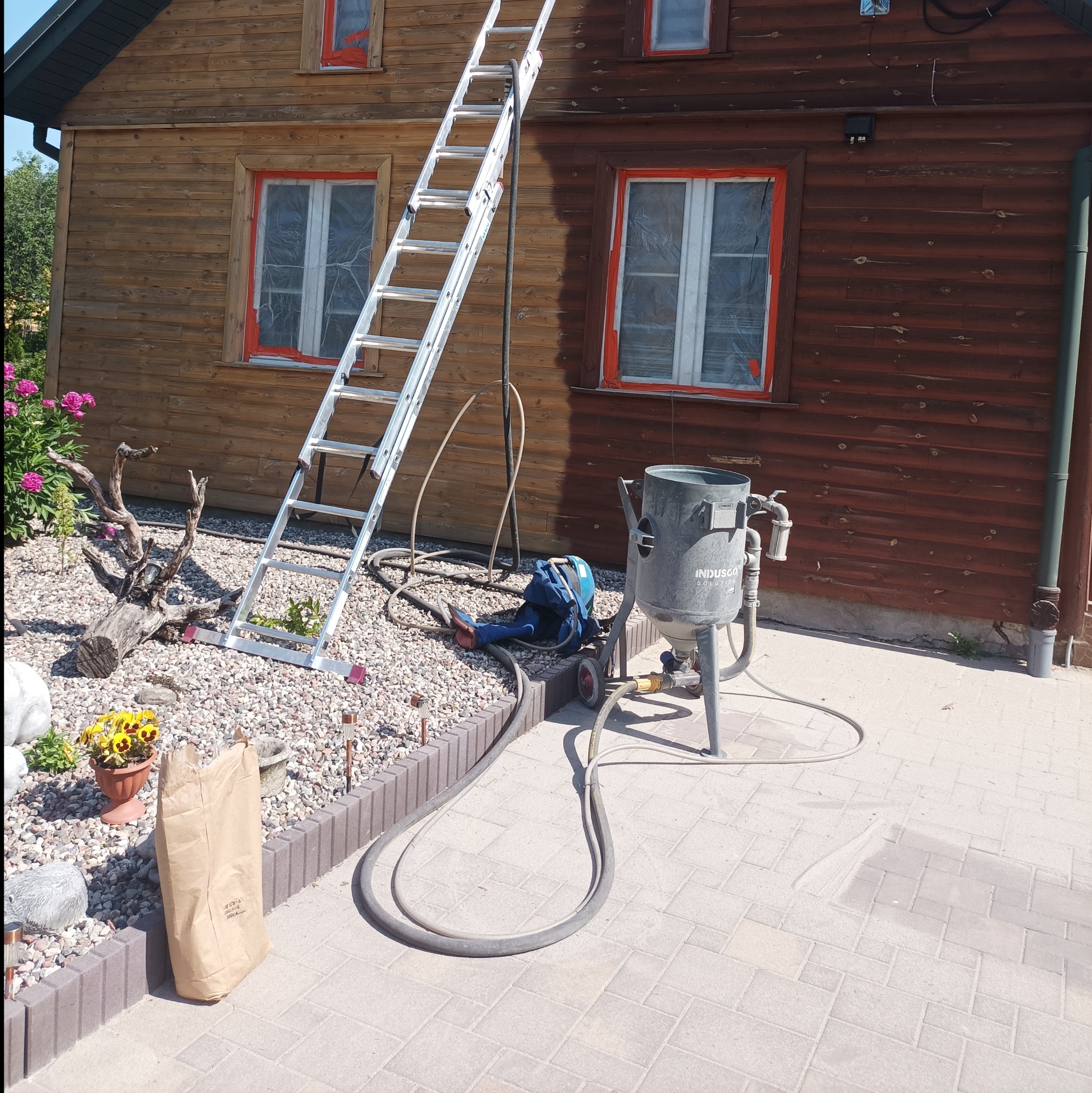 Drewniany dom z zabezpieczonymi oknami, drabina oparta o ścianę, sprzęt do obróbki strumieniowo-ściernej, torba z materiałem ściernym obok kwiatów.