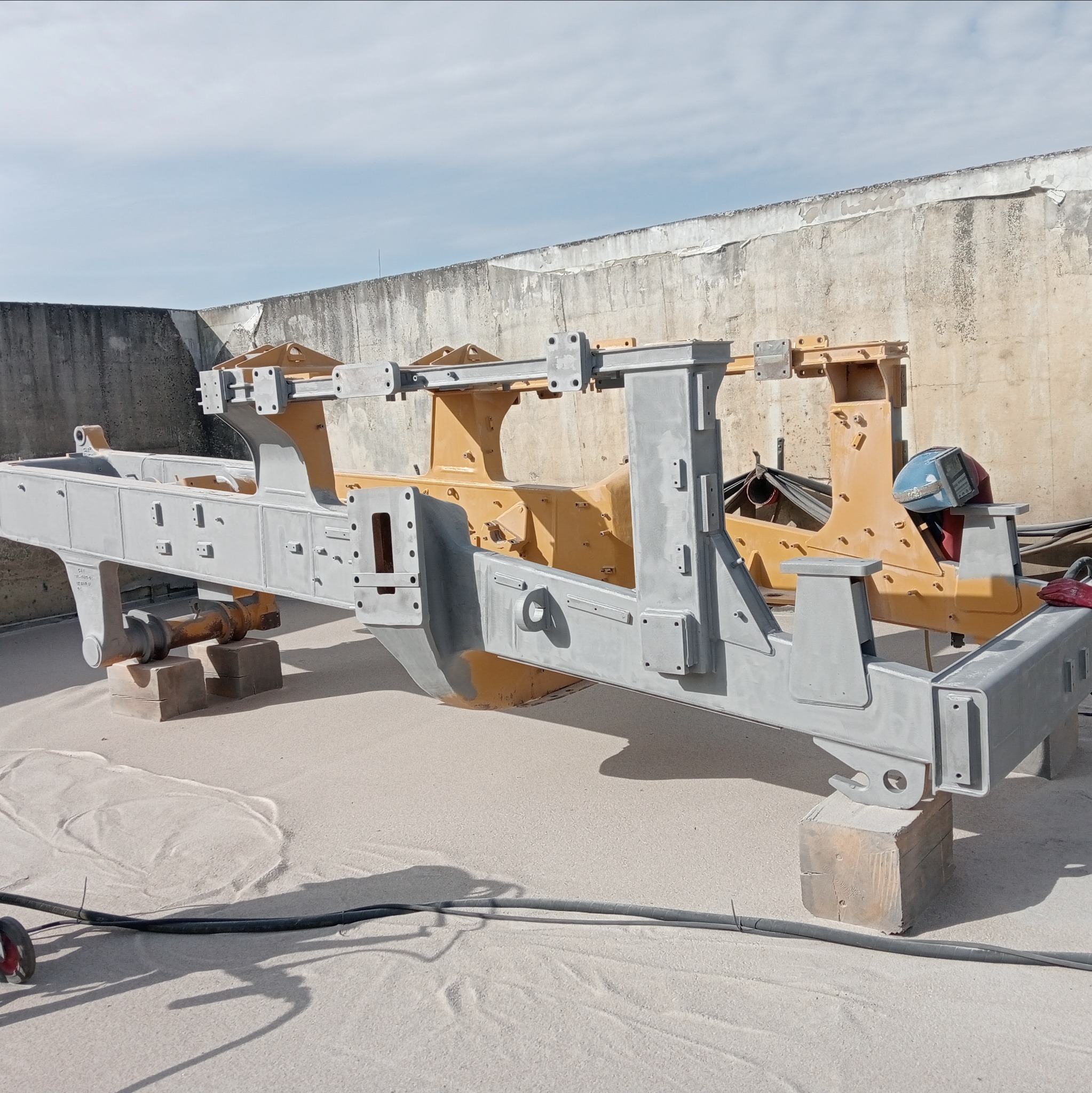 Metalowa konstrukcja przemysłowa, częściowo pomalowana szarą farbą, oparta na drewnianych klockach, z widocznymi śladami wcześniejszej, żółtej warstwy. Na konstrukcji leży niebieska maska...