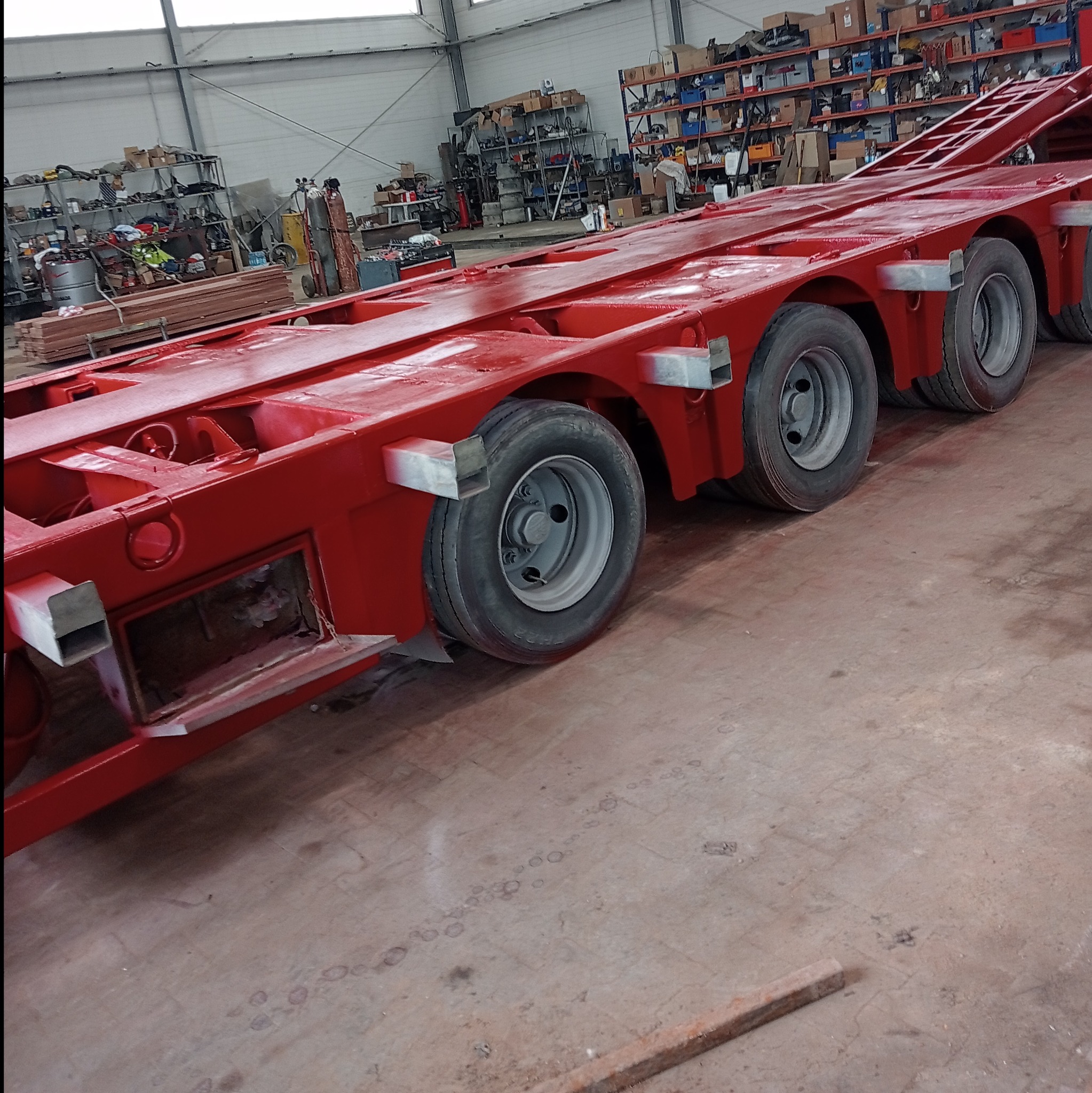 Czerwona, pomalowana rama naczepy z kołami w hali warsztatowej, ze śladami niedomalowań i zaciekami farby, widoczne półki z narzędziami w tle.