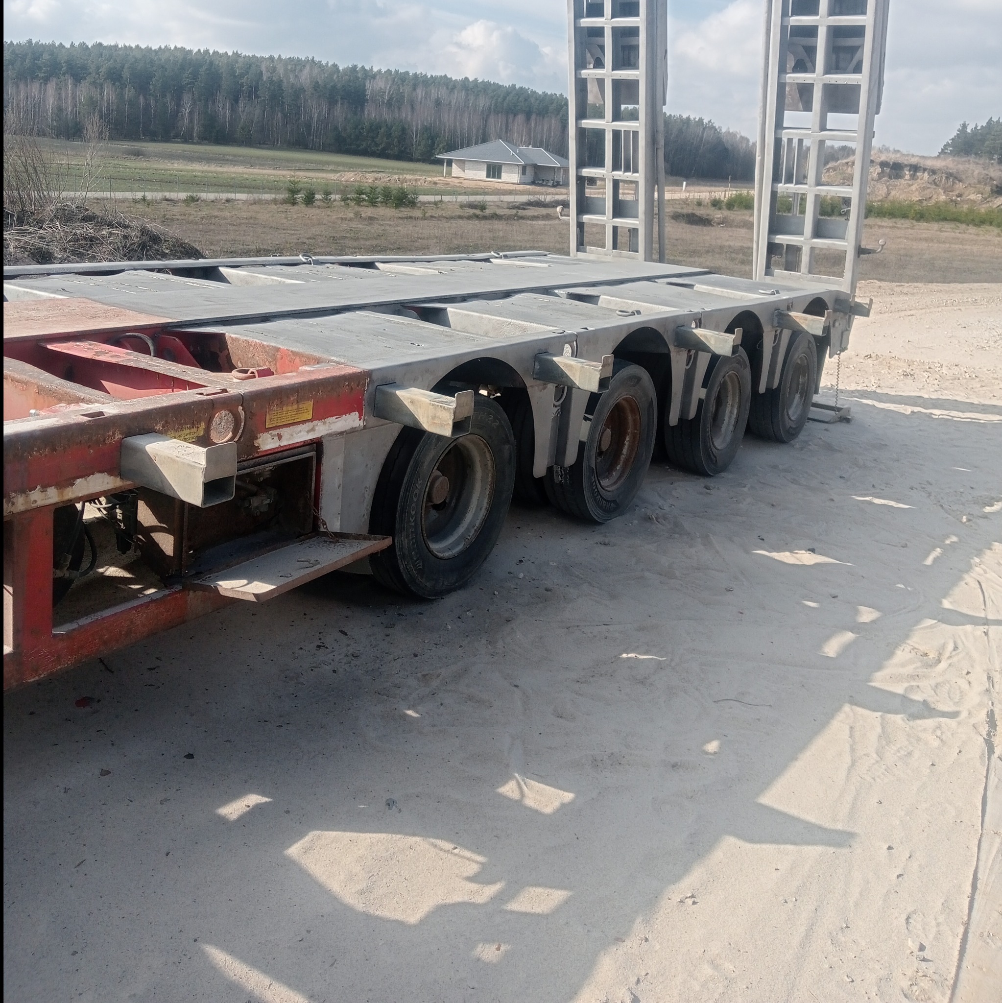 Częściowo rdzawy, wieloosiowy podnośnik hydrauliczny na piasku, z widocznym fragmentem lasu i domu w tle.