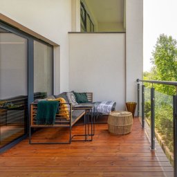 Eindom - nieruchomości - Łódź - Nowoczesny balkon z drewnianą podłogą, szklaną balustradą i meblami wypoczynkowymi w odcieniach szarości i zieleni, z widokiem na zieleń.