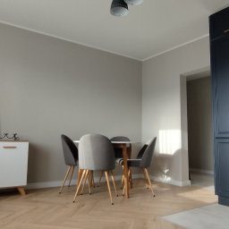 EINDOM SPÓŁKA Z OGRANICZONĄ ODPOWIEDZIALNOŚCIĄ - Jasne wnętrze: parkiet w jodełkę, biała komoda z rośliną i grafiką, stół z krzesłami, ciemna zabudowa kuchenna z miedzianymi uchwytami. Neutralna kolorystyka ścian.