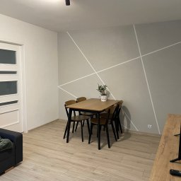 Eindom - nieruchomości - Łódź - Jasne wnętrze pokoju dziennego z drewnianym stołem i krzesłami na tle ściany z geometrycznym wzorem. Widoczny fragment sofy i telewizora.
