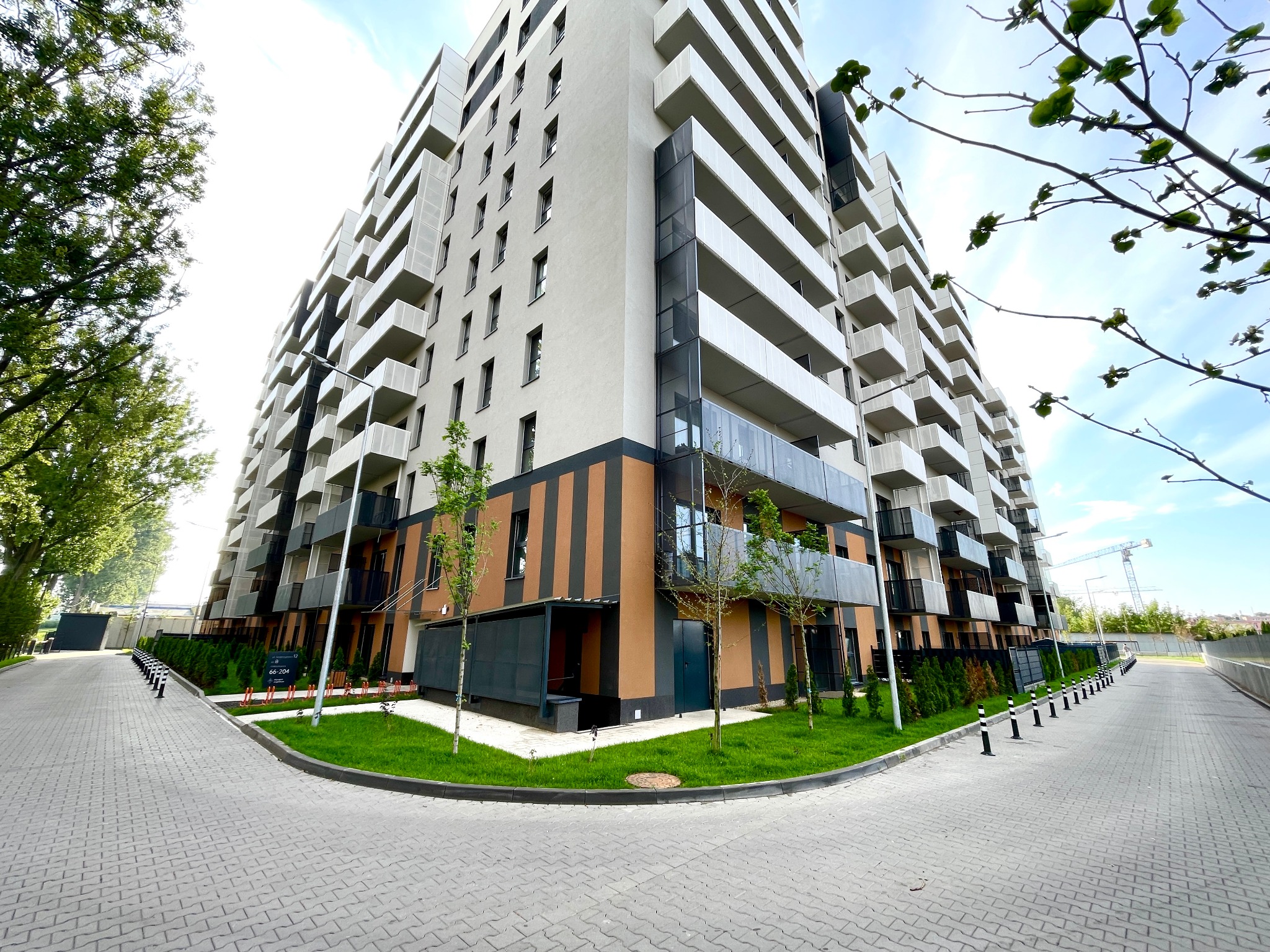 Nowoczesny apartamentowiec z balkonami, widok z poziomu chodnika. Architektura z elementami brązu i zieleni. Niebo z delikatnymi chmurami w tle.