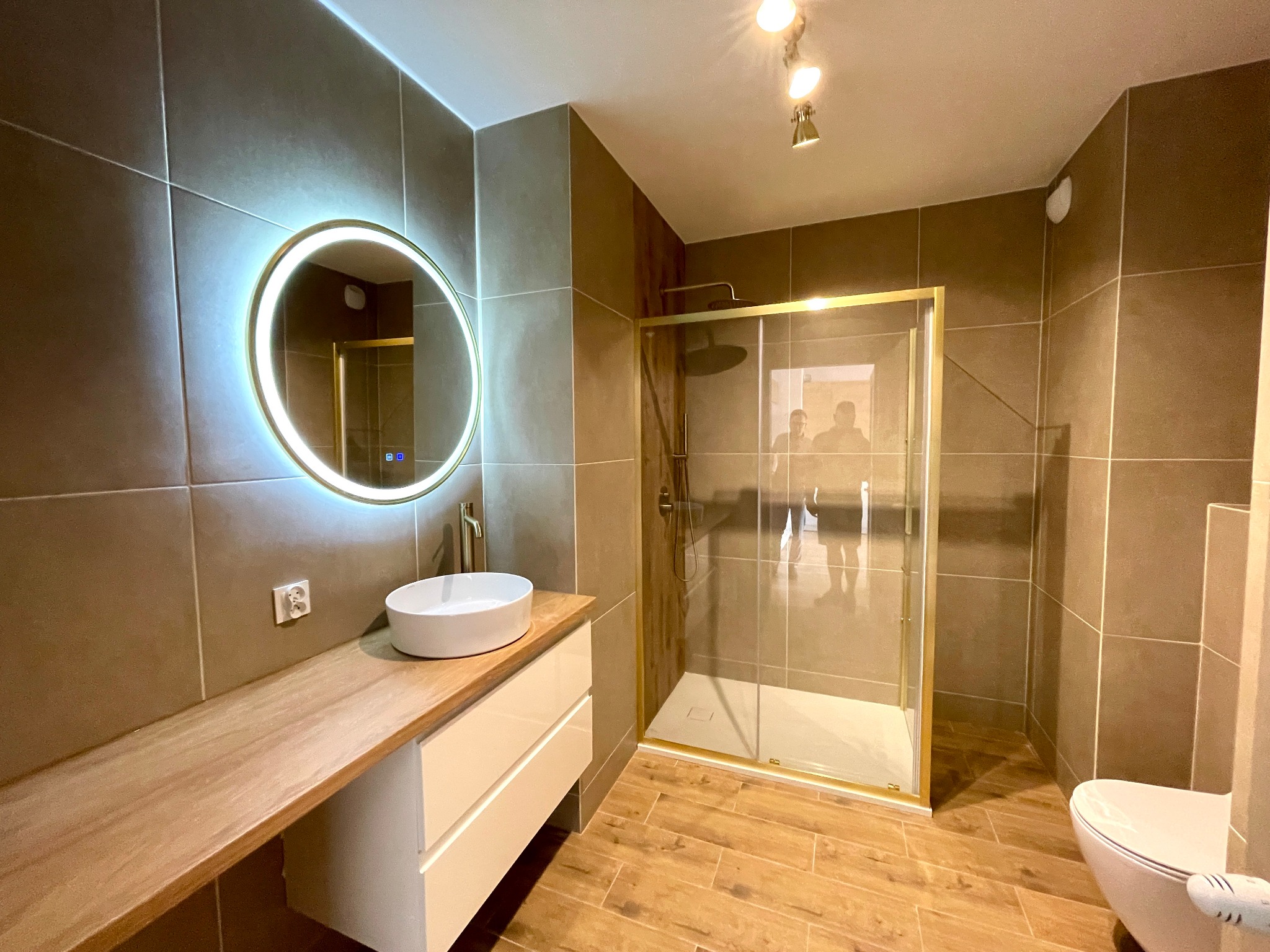 Nowoczesna łazienka z kabiną prysznicową w złotej ramie, okrągłym lustrem z podświetleniem LED i drewnianym blatem pod umywalką. Brązowe płytki na ścianach i podłodze.