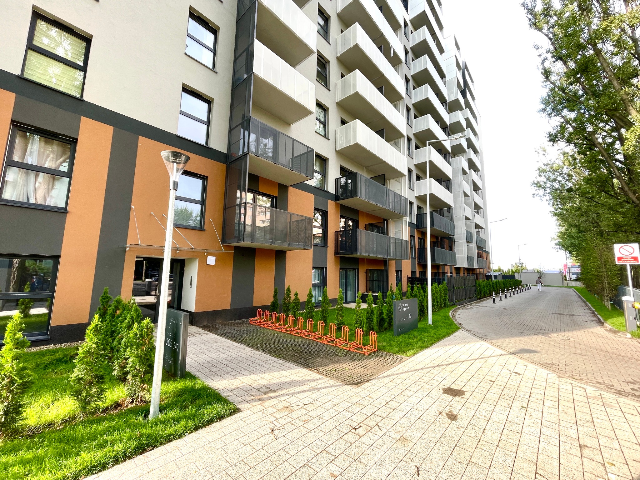 Nowoczesny budynek apartamentowy z balkonami, pomarańczowo-szarą elewacją i zagospodarowanym terenem zielonym z chodnikiem i stojakami na rowery.
