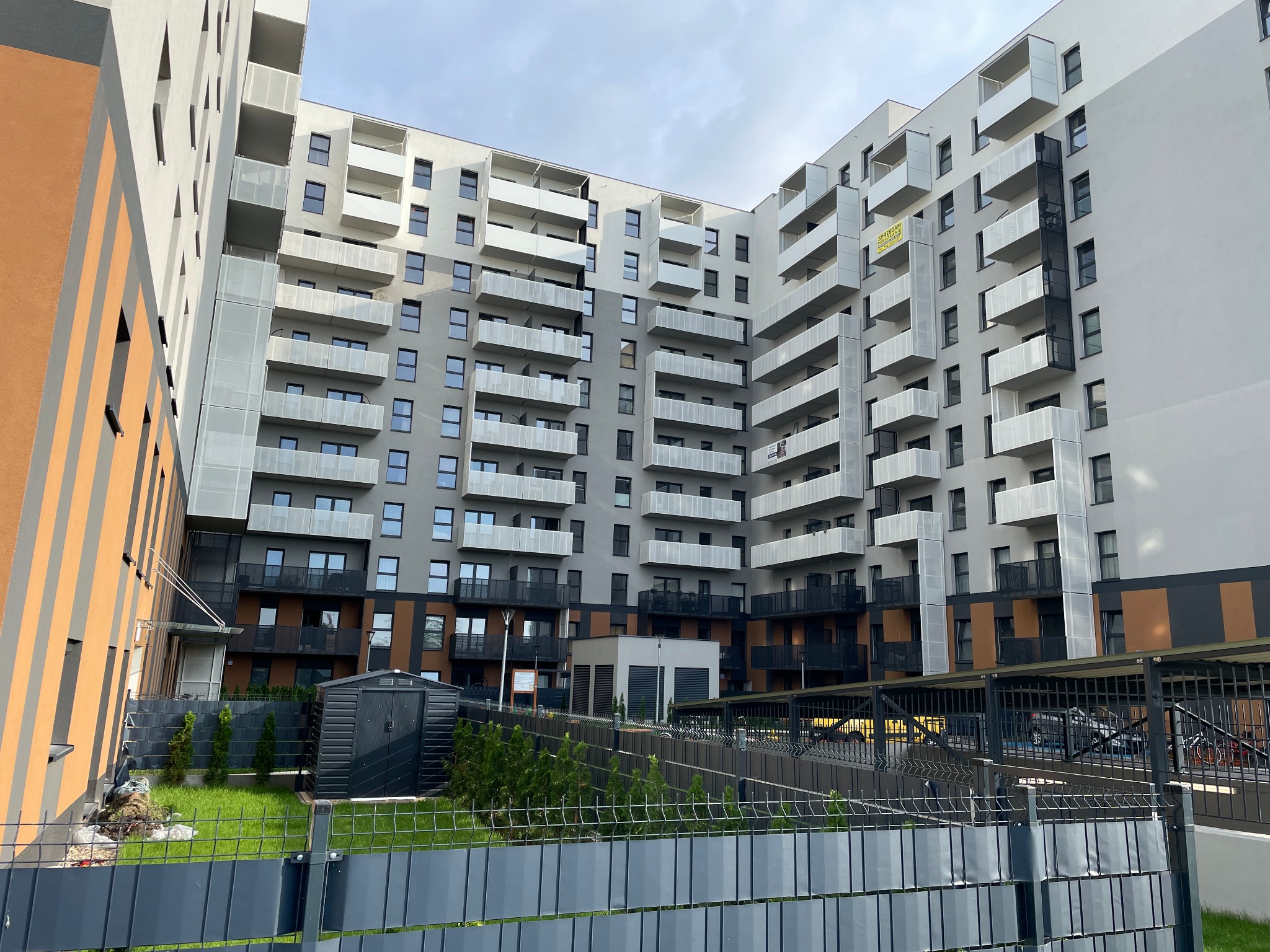 Nowoczesny apartamentowiec z białymi balkonami i szarą elewacją, widoczny zza ogrodzenia. Na jednym z balkonów widnieje baner informacyjny.