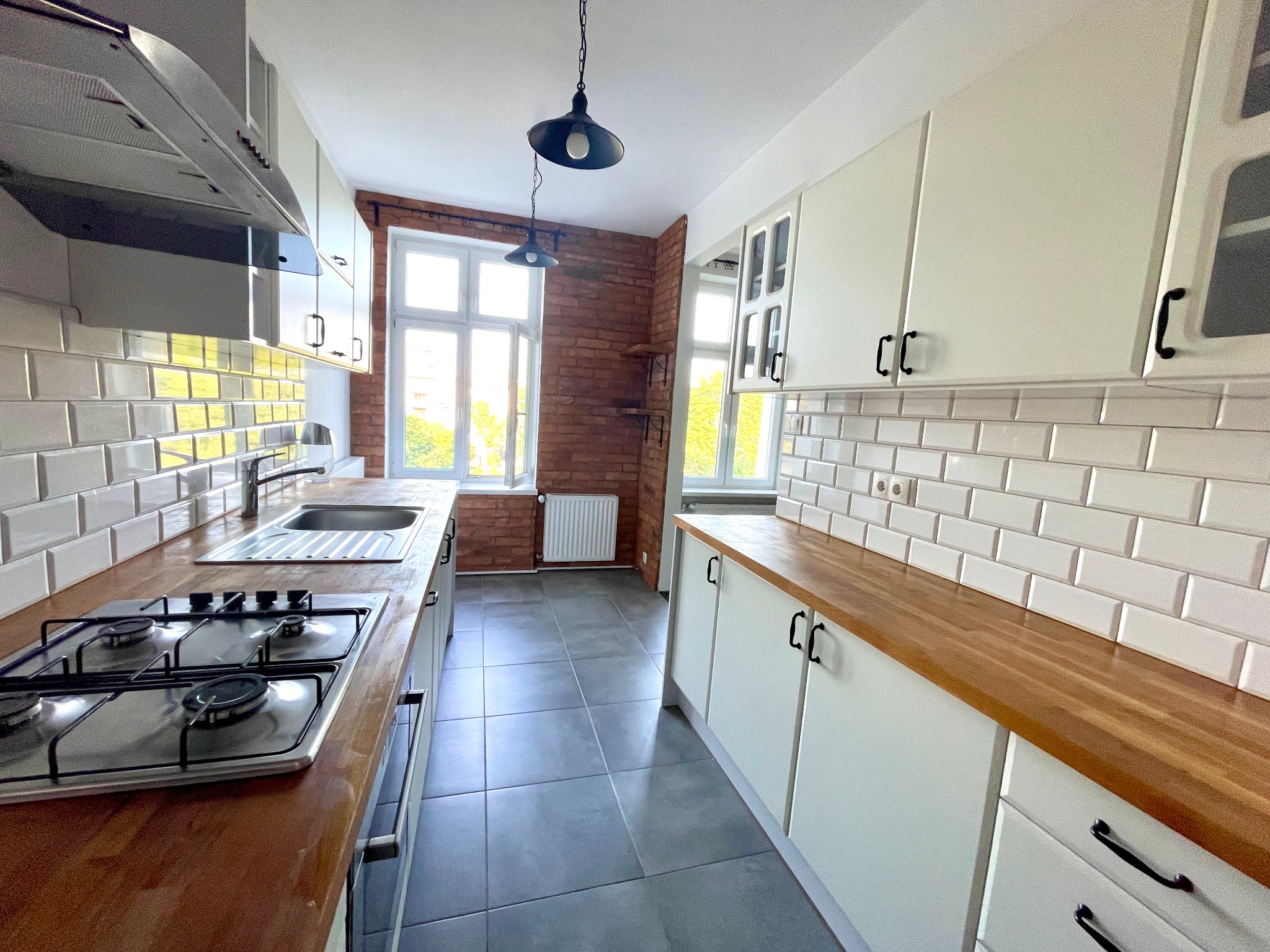 Jasna kuchnia w stylu retro: białe szafki z drewnianym blatem, ceglana ściana z oknem i szara podłoga. Widok na całe pomieszczenie.