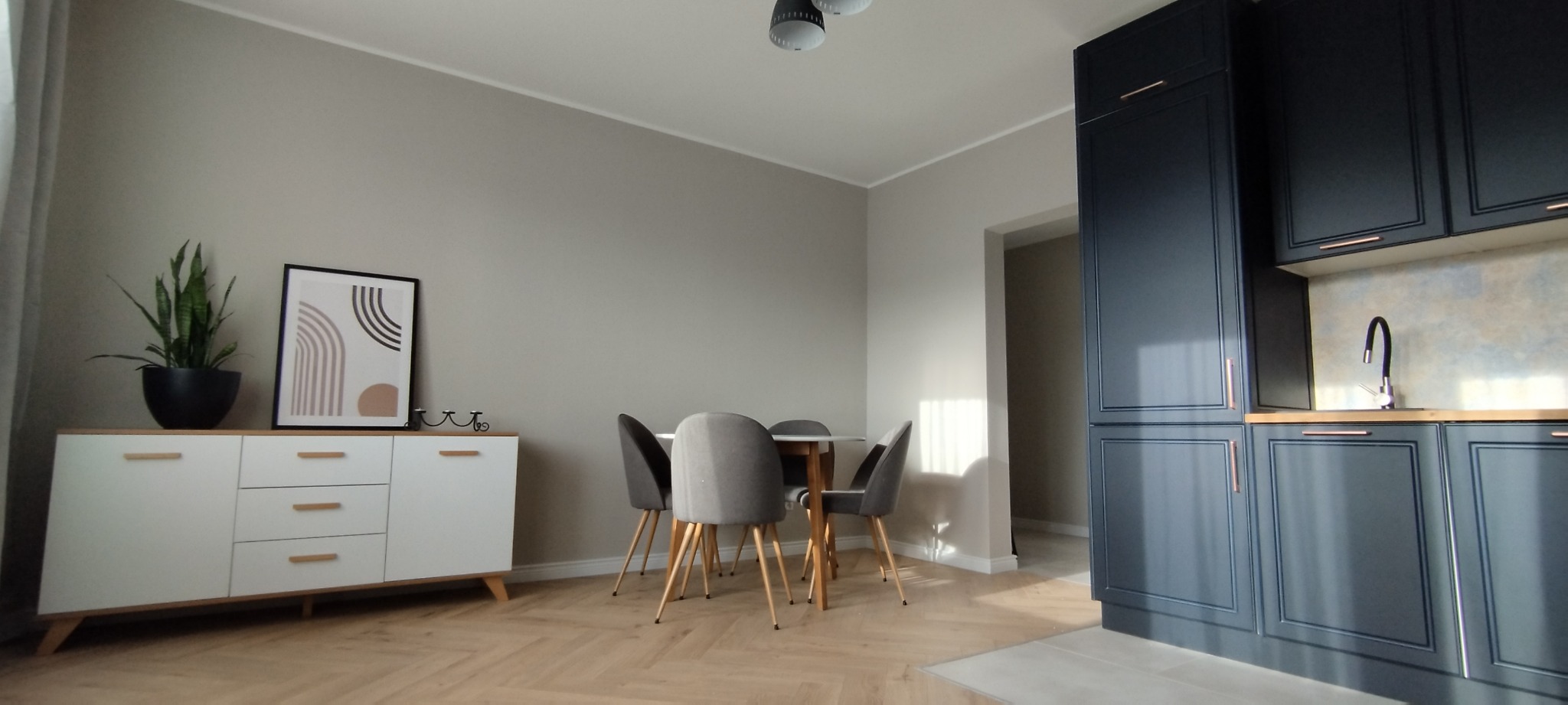 Jasne wnętrze: parkiet w jodełkę, biała komoda z rośliną i grafiką, stół z krzesłami, ciemna zabudowa kuchenna z miedzianymi uchwytami. Neutralna kolorystyka ścian.