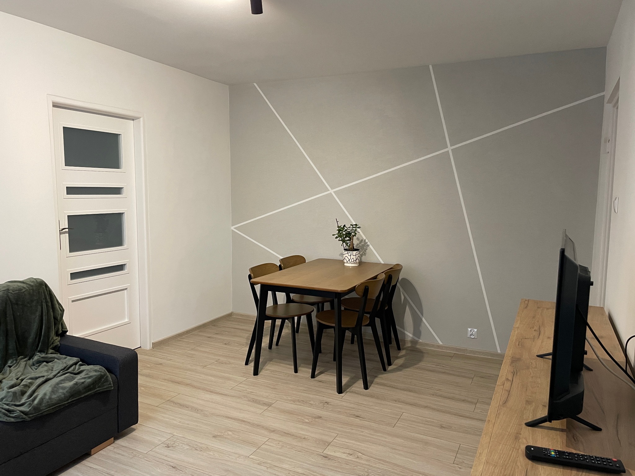 Jasne wnętrze pokoju dziennego z drewnianym stołem i krzesłami na tle ściany z geometrycznym wzorem. Widoczny fragment sofy i telewizora.