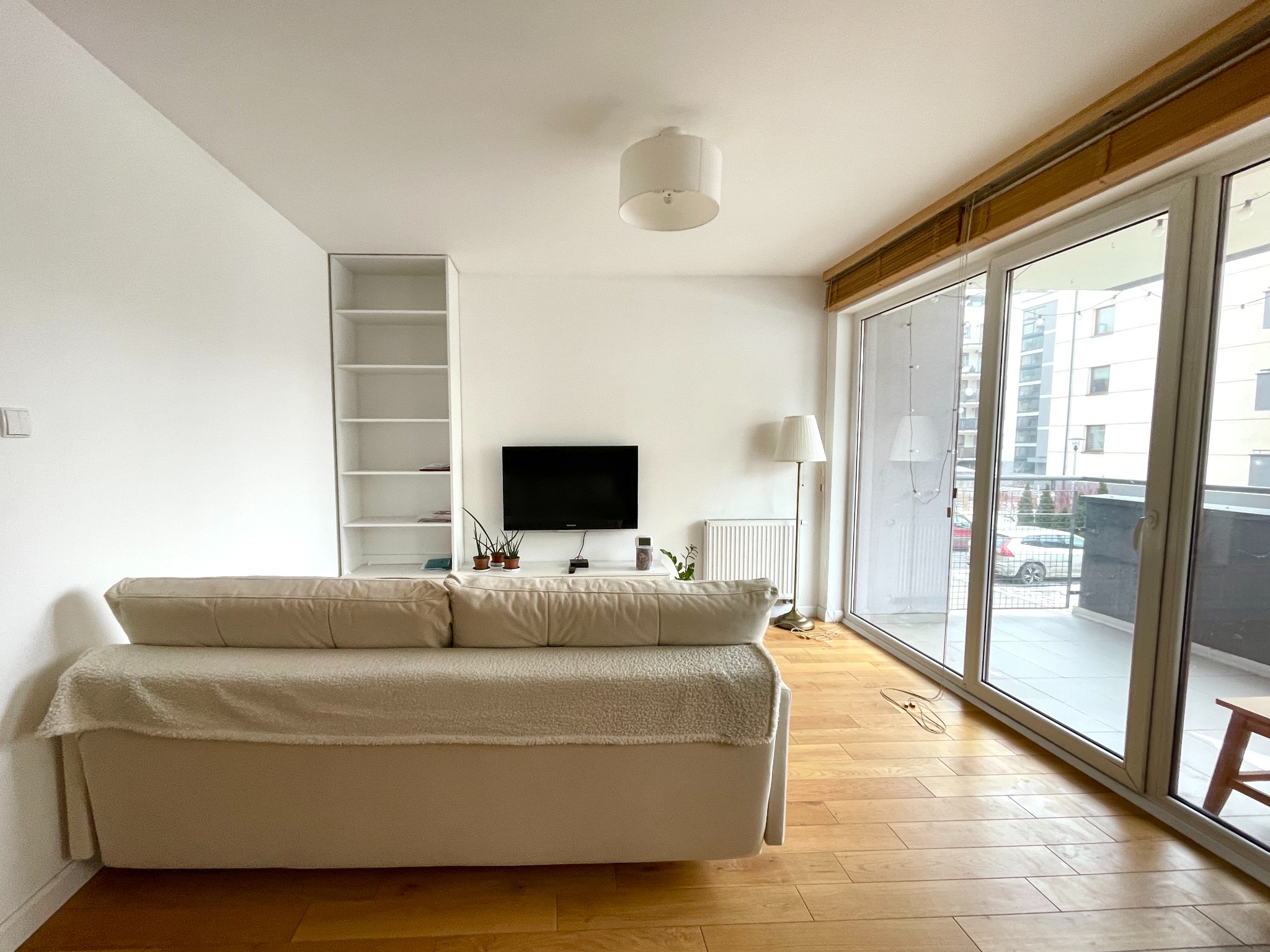 Jasne wnętrze salonu z beżową sofą, drewnianą podłogą i dużym oknem balkonowym. Widoczny regał, telewizor i lampa podłogowa. Minimalistyczny wystrój.