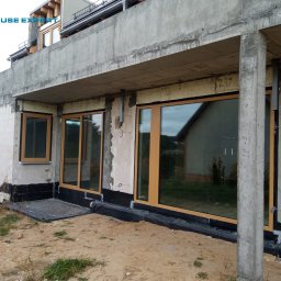 PAWEŁ KRAUSE Krause Expert - Surowy stan budynku z zamontowanymi oknami z drewnopodobną ramą i dużymi przeszkleniami, widoczne detale izolacji fundamentów i niezakończone prace wykończeniowe.