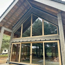 PAWEŁ KRAUSE Krause Expert - Nowoczesny dom z dużymi oknami aluminiowymi i przesuwnymi drzwiami tarasowymi, widok z zewnątrz na tle zieleni i fragmentu placu budowy.