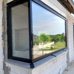 PAWEŁ KRAUSE Krause Expert - Duże okno z czarną ramą zamontowane w surowym budynku z białych bloczków, z widokiem na zielone otoczenie.