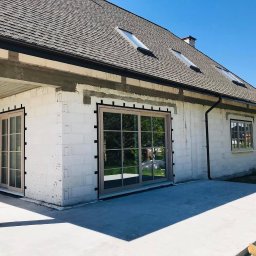 PAWEŁ KRAUSE Krause Expert - Dom w trakcie budowy z zamontowanymi oknami i drzwiami balkonowymi z jasnymi ramami, widoczne czarne kliny montażowe i świeże tynki.