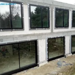 Surowy stan budynku z zamontowanymi dużymi oknami z czarnymi ramami, widoczne szczeliny wypełnione pianką montażową, w tle las.