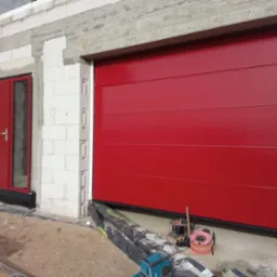 Czerwona segmentowa brama garażowa i drzwi wejściowe w surowym stanie budynku, widoczne narzędzia i materiały budowlane.