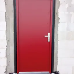 Czerwone drzwi zewnętrzne z czarną ościeżnicą zamontowane w surowym murze z betonu komórkowego, widoczna izolacja przeciwwilgociowa u dołu.