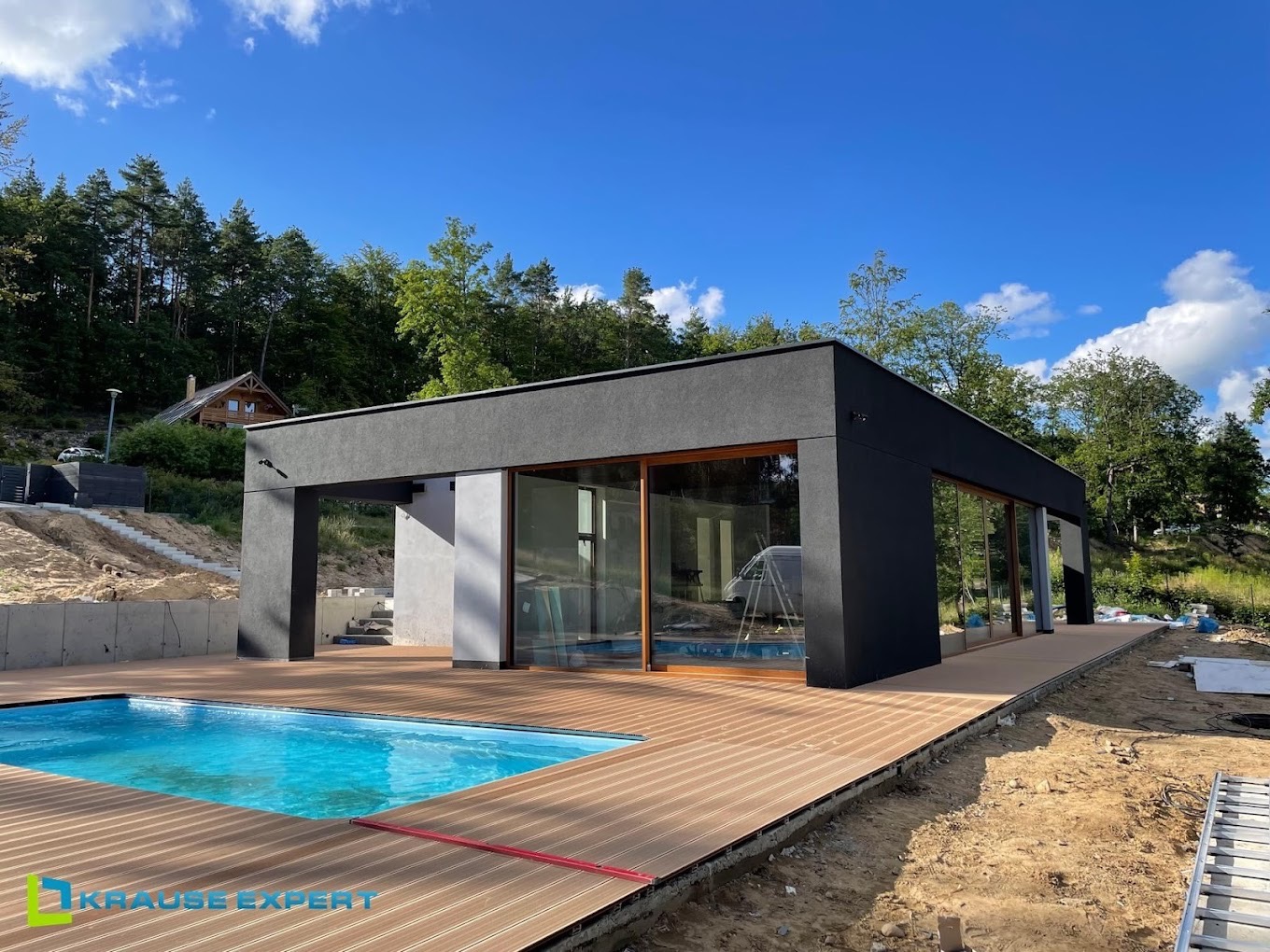 Nowoczesny, czarny budynek z dużymi przeszkleniami i drewnianym tarasem przy basenie, w tle zalesione wzgórze i błękitne niebo.