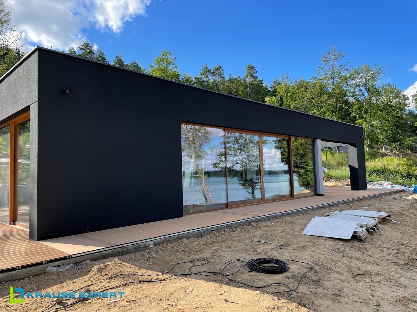 Nowoczesny, czarny dom z dużymi przesuwnymi oknami z drewnianymi ramami i tarasem z desek kompozytowych, widok na jezioro, prace wykończeniowe w toku.