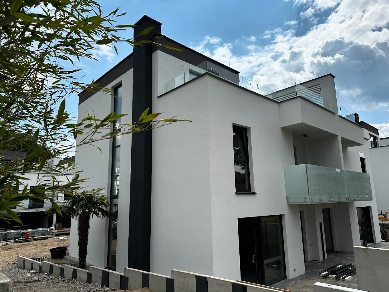 Nowoczesny, dwupiętrowy dom jednorodzinny z białymi ścianami, czarnymi akcentami i szklanymi balustradami balkonów, widoczny z perspektywy narożnej, na tle błękitnego nieba z chmurami, z palmą...