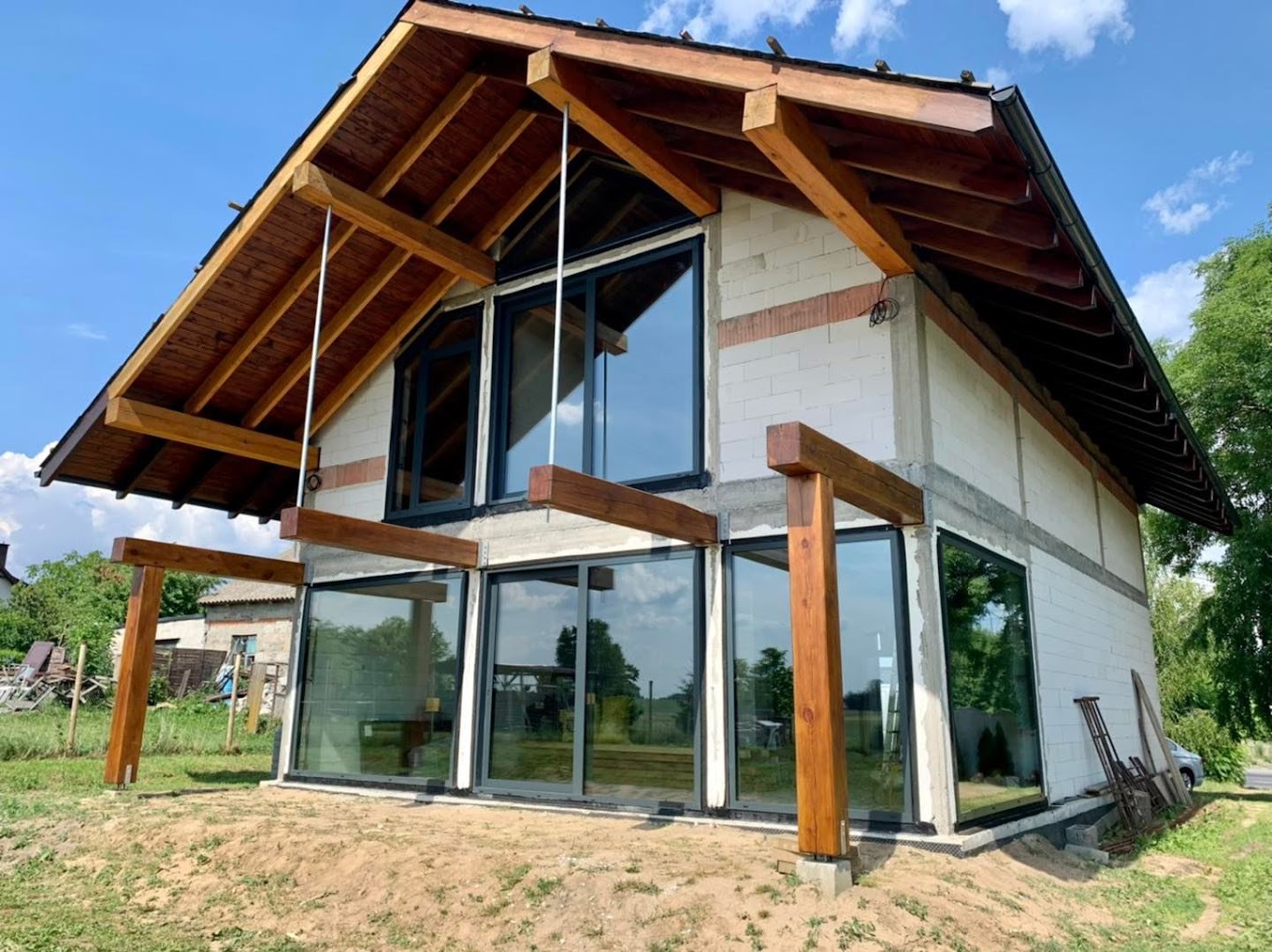 Dom w trakcie budowy z dużymi oknami aluminiowymi, widoczne drewniane elementy konstrukcyjne dachu i zadaszenia tarasu, ściany z białego bloczka.