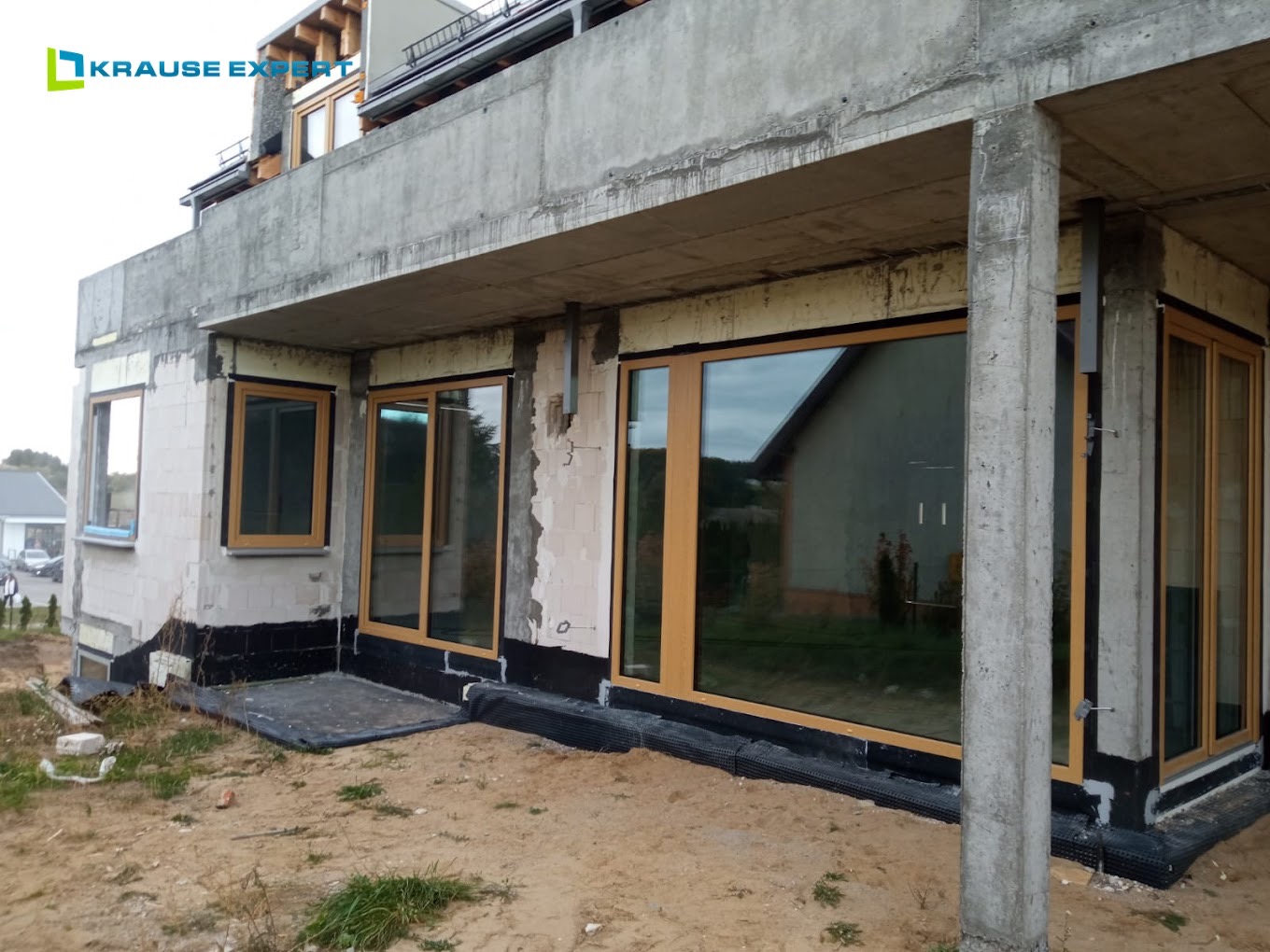Surowy stan budynku z zamontowanymi oknami z drewnopodobną ramą i dużymi przeszkleniami, widoczne detale izolacji fundamentów i niezakończone prace wykończeniowe.