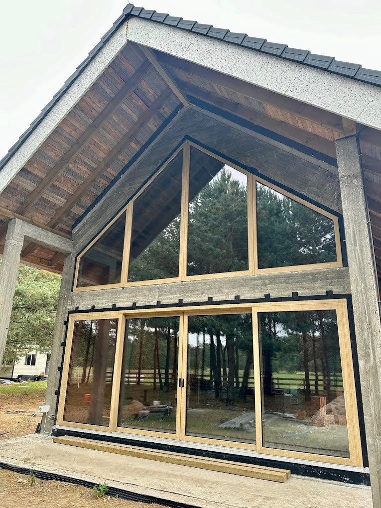 Nowoczesny dom z dużymi oknami aluminiowymi i przesuwnymi drzwiami tarasowymi, widok z zewnątrz na tle zieleni i fragmentu placu budowy.