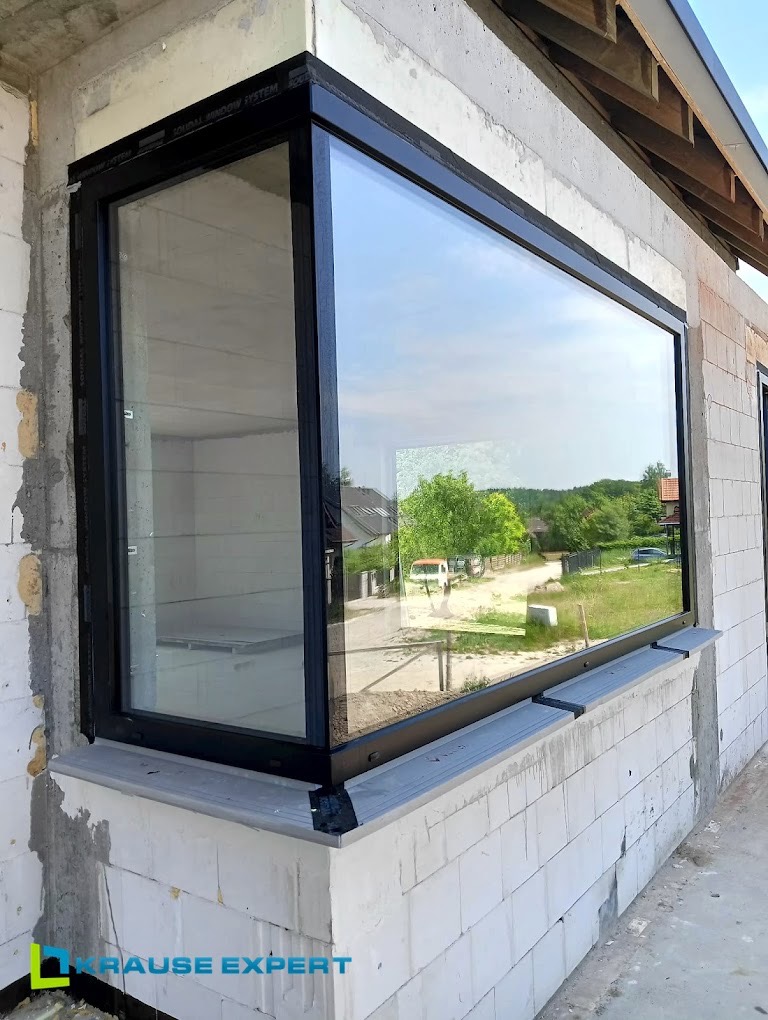 Duże okno z czarną ramą zamontowane w surowym budynku z białych bloczków, z widokiem na zielone otoczenie.