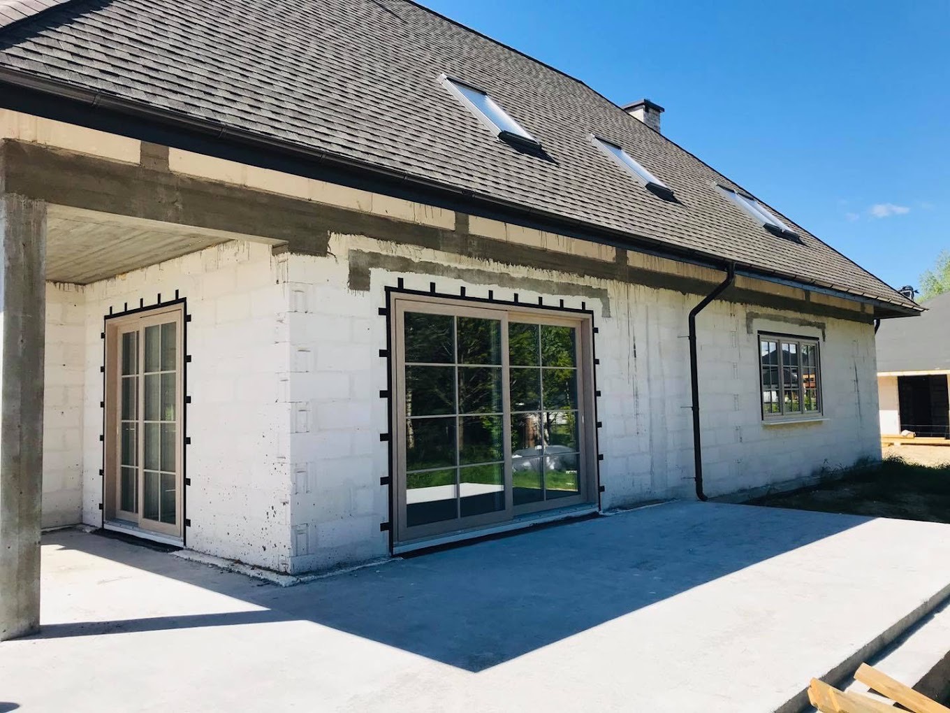 Dom w trakcie budowy z zamontowanymi oknami i drzwiami balkonowymi z jasnymi ramami, widoczne czarne kliny montażowe i świeże tynki.