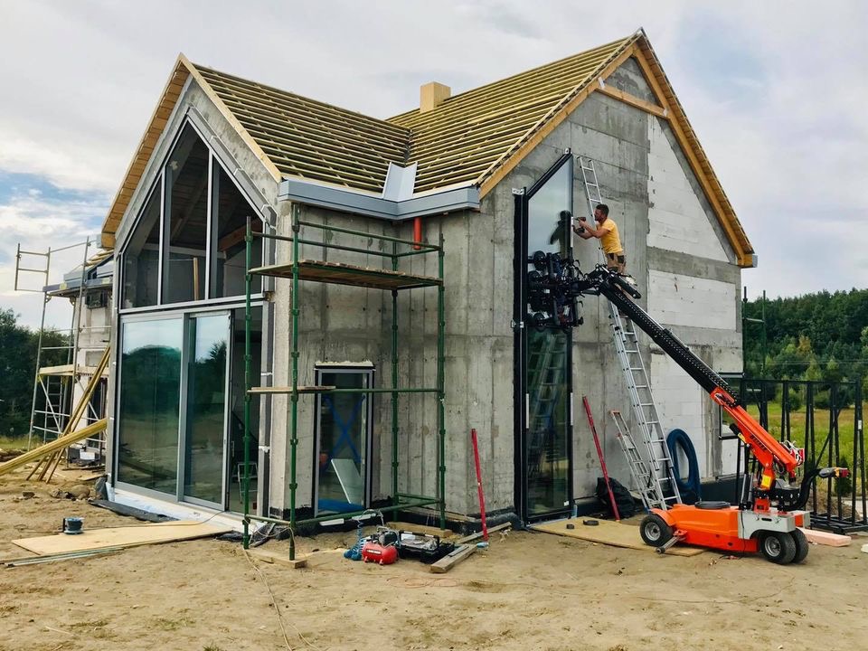 Montaż dużego, panoramicznego okna w surowym stanie domu jednorodzinnego przy użyciu specjalistycznego dźwigu próżniowego i rusztowań.