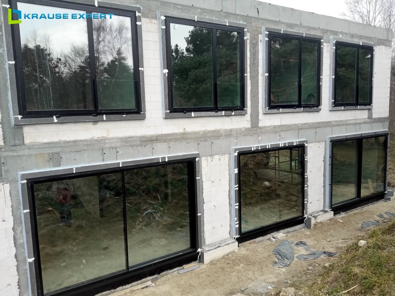 Surowy stan budynku z zamontowanymi dużymi oknami z czarnymi ramami, widoczne szczeliny wypełnione pianką montażową, w tle las.