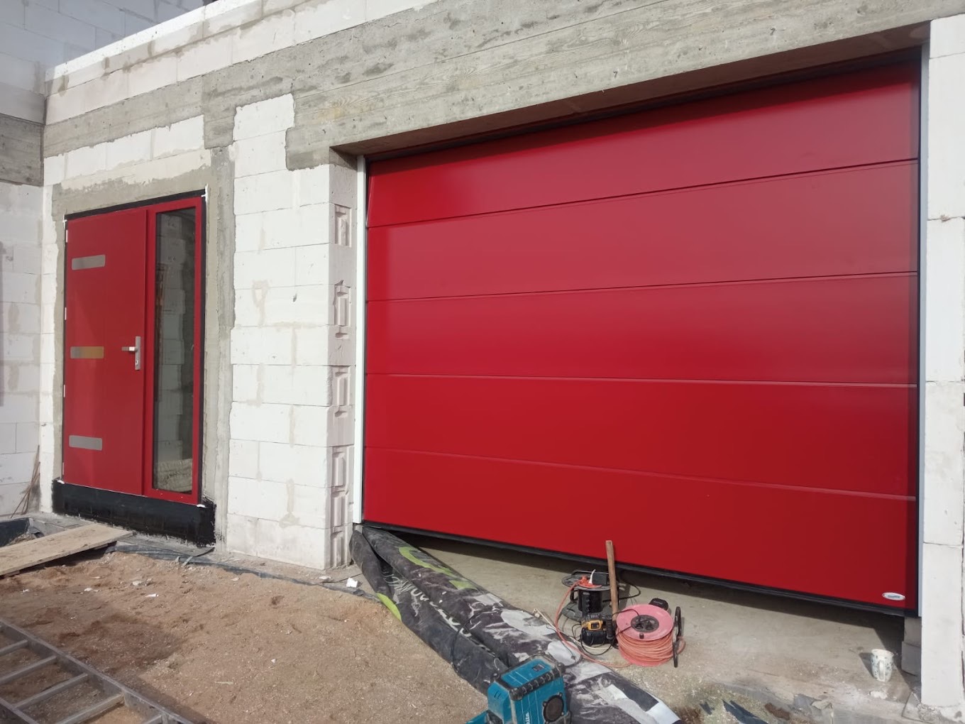 Czerwona segmentowa brama garażowa i drzwi wejściowe w surowym stanie budynku, widoczne narzędzia i materiały budowlane.