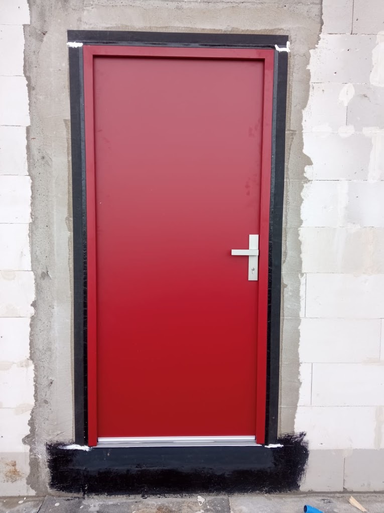 Czerwone drzwi zewnętrzne z czarną ościeżnicą zamontowane w surowym murze z betonu komórkowego, widoczna izolacja przeciwwilgociowa u dołu.