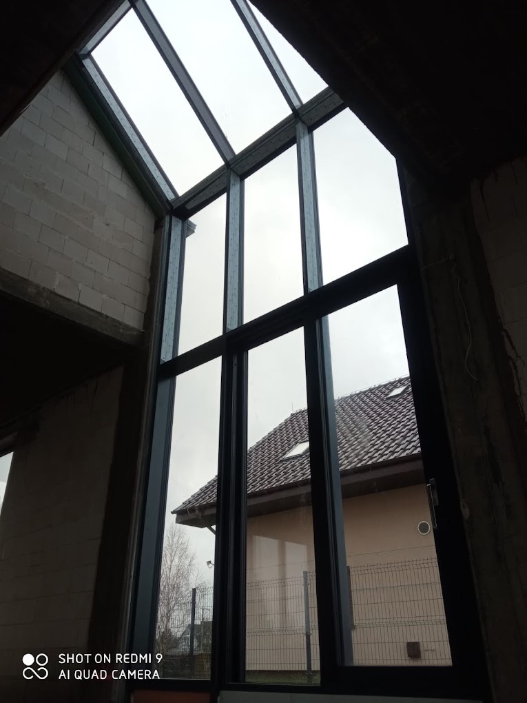 Wysokie przeszklenie w surowym stanie budynku, widok na dom z brązowym dachem przez okno z czarnymi ramami.