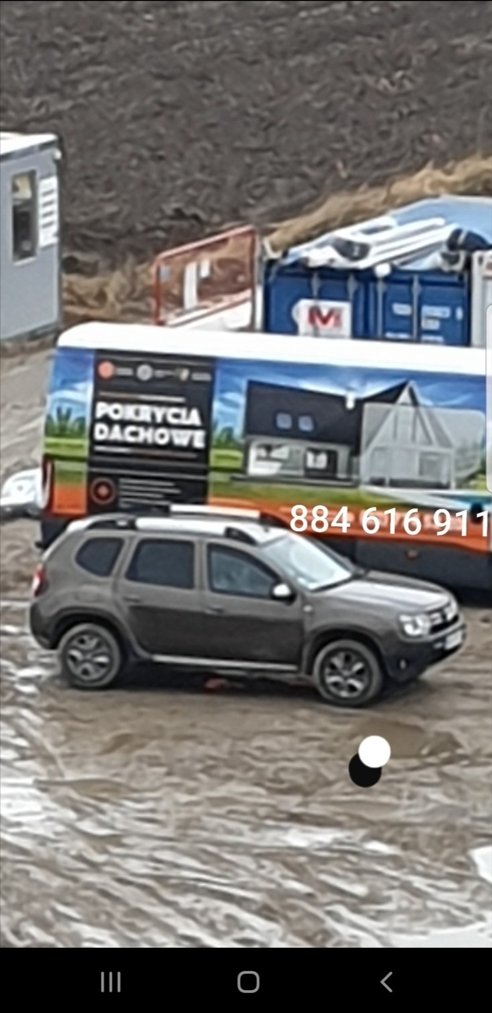 Szary samochód osobowy na błotnistym terenie, w tle przyczepa z reklamą firmy dekarskiej z numerem telefonu.