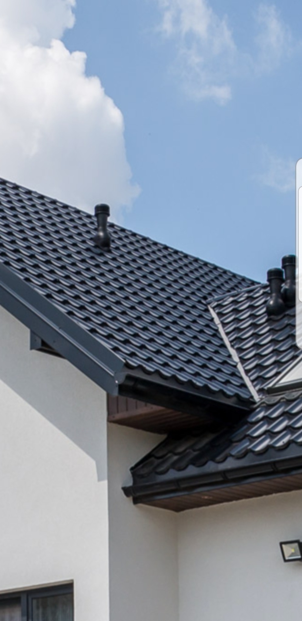 Fragment domu z czarnym, metalowym pokryciem dachowym, widoczne kominki wentylacyjne i rynny w kolorze dachu, na tle błękitnego nieba z chmurami.