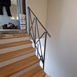 DMYTRO MILCHAKOVSKYI - Drewniane schody z szarą krawędzią, metalowa balustrada o geometrycznym wzorze, w tle widoczne wiaderka z farbą i narzędzia malarskie.