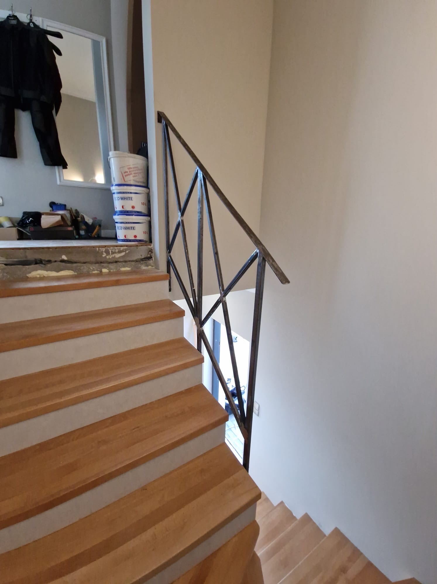 Drewniane schody z szarą krawędzią, metalowa balustrada o geometrycznym wzorze, w tle widoczne wiaderka z farbą i narzędzia malarskie.