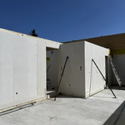 Wznoszenie ścian domu szkieletowego z prefabrykatów betonowych, widoczne podpory stabilizujące i drabina oparta o ścianę z żółtą folią ochronną na tle błękitnego nieba.