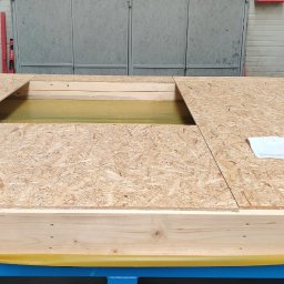 DEWERIS HOLDING PROSTA SPÓŁKA AKCYJNA - Fragment konstrukcji podłogi z płyty OSB z otworem, umieszczonej na niebieskiej ramie, zabezpieczonej żółtą folią, z widocznym planem na papierze.