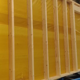 DEWERIS HOLDING PROSTA SPÓŁKA AKCYJNA - Fragment ściany szkieletowej z żółtą membraną wewnątrz ramy, widoczna konstrukcja drewniana, przygotowana do transportu na niebieskim wózku transportowym w hali produkcyjnej.