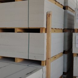 DEWERIS HOLDING PROSTA SPÓŁKA AKCYJNA - Składowane płyty gipsowo-kartonowe na paletach, zabezpieczone zielonymi taśmami, z widocznymi etykietami producenta.