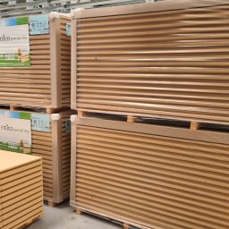 DEWERIS HOLDING PROSTA SPÓŁKA AKCYJNA - Palety z płytami izolacyjnymi Steico Special Dry, owinięte folią, składowane w magazynie, widoczna butelka wody na jednej z palet.