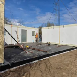 Montaż modułowej konstrukcji domu: robotnicy w kamizelkach montują ściany na przygotowanym fundamencie, widoczne narzędzia i elementy konstrukcyjne.