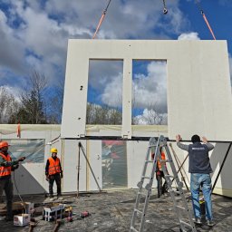 DEWERIS HOLDING PROSTA SPÓŁKA AKCYJNA - Montaż segmentu domu modułowego z oknami przez ekipę budowlaną. Element podnoszony dźwigiem, pracownicy w kaskach i kamizelkach. Widoczne narzędzia i drabina.