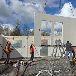 DEWERIS HOLDING PROSTA SPÓŁKA AKCYJNA - Montaż ściany z dwoma oknami na budowie domu modułowego. Pracownicy w kaskach i kamizelkach, drabina, narzędzia. Jasne, pochmurne niebo w tle.