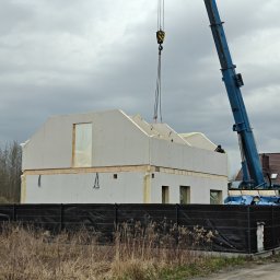 DEWERIS HOLDING PROSTA SPÓŁKA AKCYJNA - Montaż segmentu domu szkieletowego za pomocą dźwigu na tle pochmurnego nieba. Widoczne elementy konstrukcyjne i zabezpieczenia.